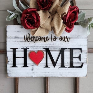 May include: A rustic wooden sign with the words "Welcome to our HOME" in black lettering. A red heart is in the center of the "O". The sign is decorated with burgundy roses, burlap, and faux greenery. The sign is mounted on a decorative window frame.