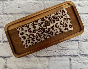 May include: A brown and beige leopard print headband with a knotted design. The headband is on a wooden tray.