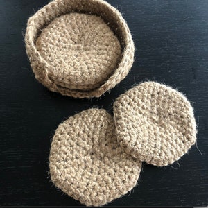 May include: Three round, crocheted jute coasters in a natural brown color. The coasters are stacked on top of each other.