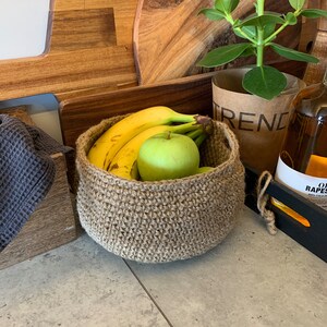 May include: A brown crocheted basket filled with bananas and a green apple. The basket is sitting on a gray countertop with a wooden cutting board and a potted plant in the background.