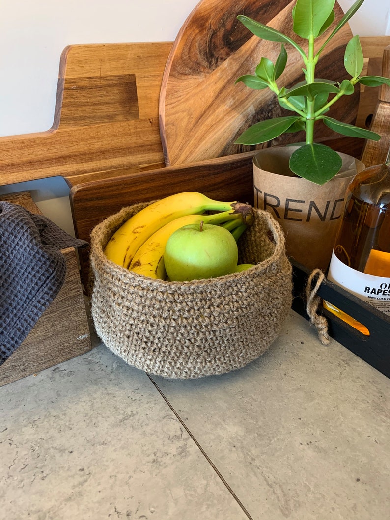 May include: A brown crocheted basket filled with bananas and a green apple. The basket is sitting on a gray countertop with a wooden cutting board and a potted plant in the background.