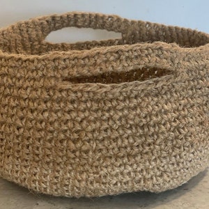May include: A natural brown crocheted basket with a single handle. The basket is made of a woven material and has a round shape.