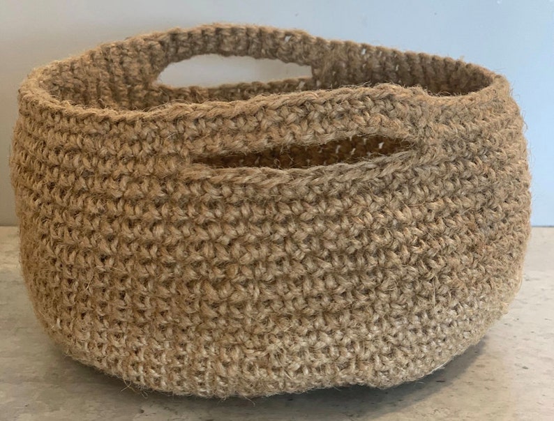 May include: A natural brown crocheted basket with a single handle. The basket is made of a woven material and has a round shape.