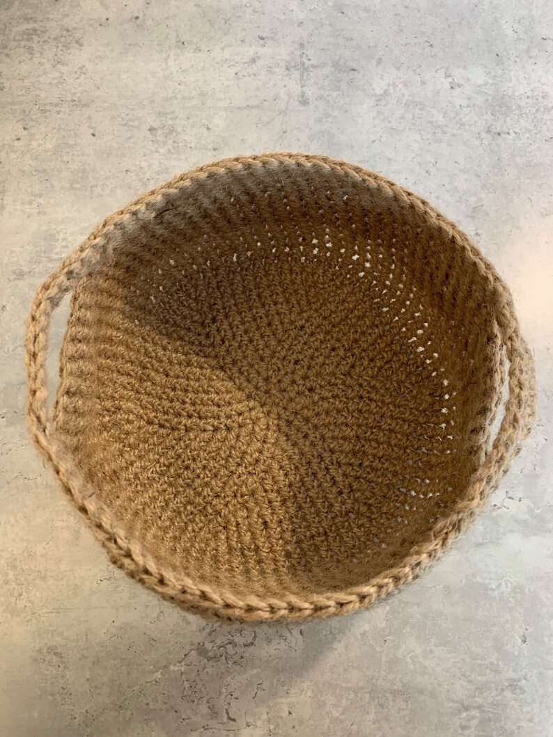 May include: A round, tan, crocheted basket with two handles. The basket is made of natural fibers and has a woven texture.