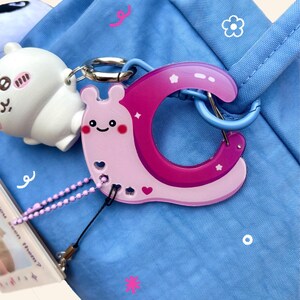May include: A light pink and magenta snail-shaped keychain with a smiling face and heart details, attached to a blue bag. A white cartoon character and a small card with text are also visible.