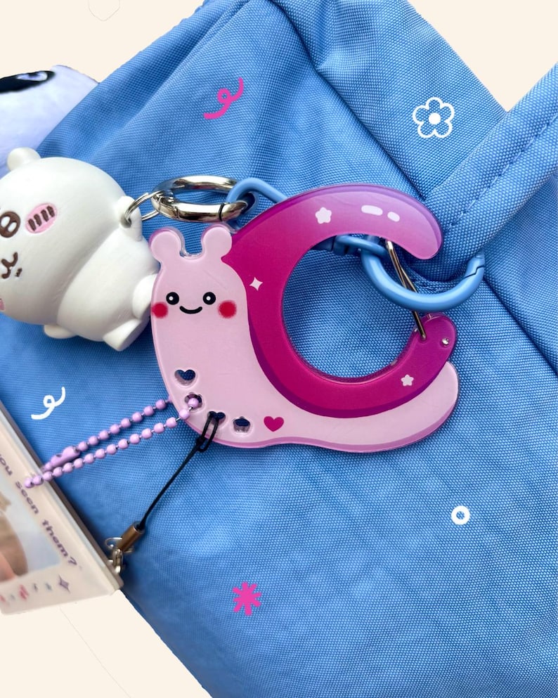 May include: A light pink and magenta snail-shaped keychain with a smiling face and heart details, attached to a blue bag. A white cartoon character and a small card with text are also visible.
