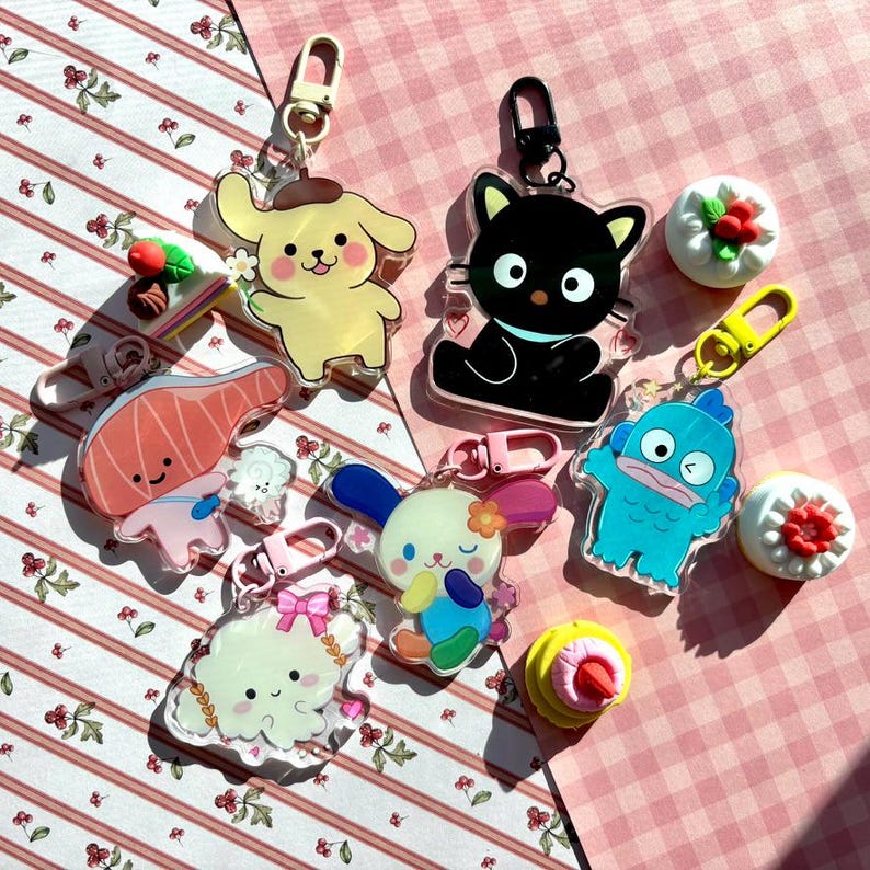 May include: A collection of colorful acrylic keychains featuring various cartoon characters and food-themed designs. The keychains include a yellow dog, a black cat, a blue monster, a bunny, and a piece of salmon. Some keychains have gold-colored clasps.