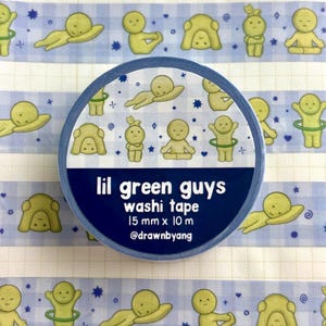 May include: A roll of washi tape featuring a light blue and white checkered pattern with small green cartoon figures in various poses, stars, and hearts. The label reads lil green guys washi tape 15 mm x 10 m @drawnbyang in white text.