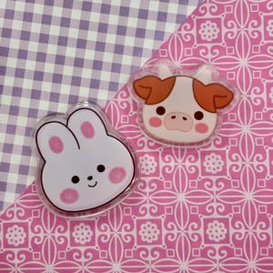 May include: Two clear acrylic pins with cartoon animal faces. One is a white bunny with pink accents, the other is a cow with brown and pink details. The pins are on a pink and purple patterned background.
