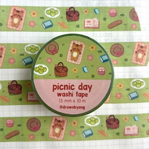 May include: A roll of green washi tape featuring a repeating pattern of picnic-themed illustrations including cats on blankets, baskets, grapes, books, and flowers. The label reads picnic day washi tape 15 mm x 10 m @drawnbyang.