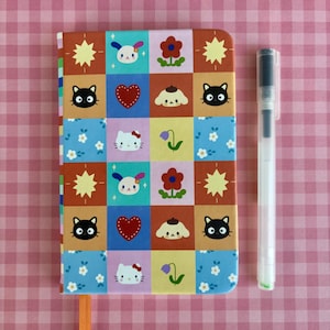 Op de afbeelding: Een kleurrijk notitieboek met een patroon op de kaft met stripfiguren, bloemen en sterren. Een oranje lint als bladwijzer is zichtbaar. Een doorzichtige pen met zwarte dop ligt naast het notitieboek.