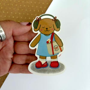 May include: A sticker featuring a cartoon dog wearing headphones and a blue dress, carrying a Trader Joe's tote bag with flowers. The dog is wearing red clogs and white socks. The sticker is on a gray oval.