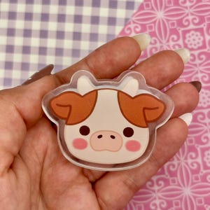 May include: A clear acrylic cow-shaped charm with a cartoon design. The cow has a white face with brown ears and spots, pink cheeks, and black eyes. The charm is held in a hand against a patterned background.