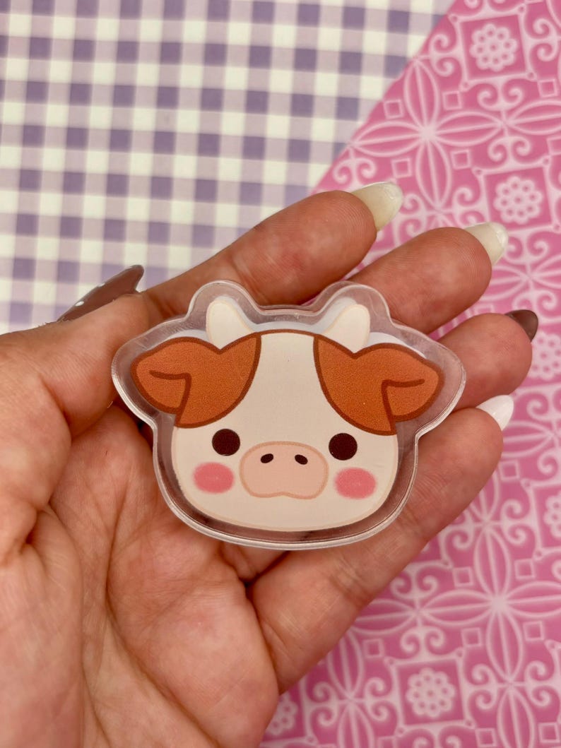 May include: A clear acrylic cow-shaped charm with a cartoon design. The cow has a white face with brown ears and spots, pink cheeks, and black eyes. The charm is held in a hand against a patterned background.