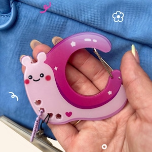 May include: A pink and purple carabiner clip in the shape of a snail with a smiling face, hearts, and stars. The clip is held in a hand against a blue background.