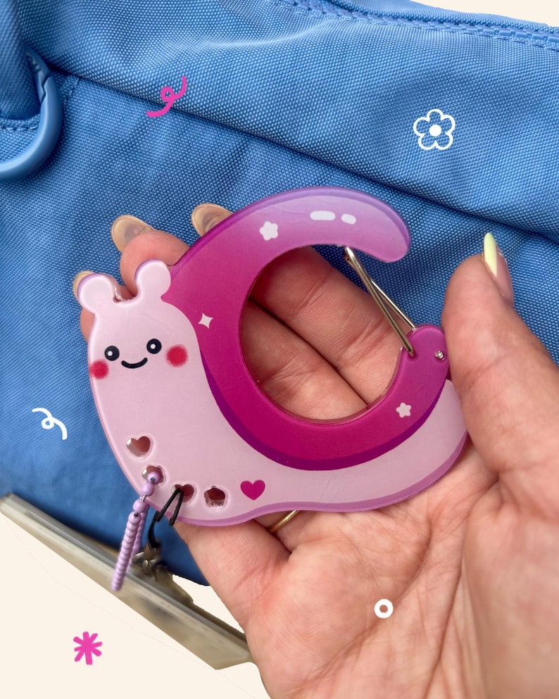 May include: A pink and purple carabiner clip in the shape of a snail with a smiling face, hearts, and stars. The clip is held in a hand against a blue background.