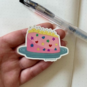 May include: A pink sticker of a birthday cake with yellow frosting, candles, and colorful star and heart decorations. The sticker is on a blue plate and has the text "@drawnbyang". A clear pen is in the background.