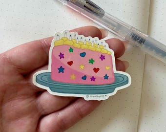 Peut inclure: Un autocollant rose en forme de gâteau d'anniversaire avec un glaçage jaune, des bougies et des décorations en forme d'étoiles et de cœurs colorés. L'autocollant est sur une assiette bleue et porte le texte "@drawnbyang". Un stylo transparent est en arrière-plan.