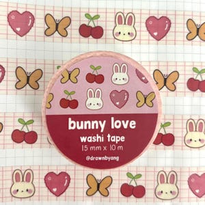May include: A roll of washi tape with a pink label featuring the text bunny love washi tape 15 mm x 10 m @drawnbyang. The tape pattern includes repeating illustrations of white bunnies, red cherries, yellow butterflies, and pink hearts on a grid.