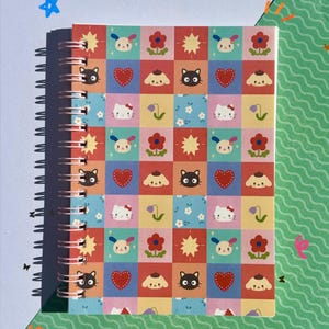 May include: A spiral-bound notebook featuring a colorful grid pattern of Sanrio characters including Hello Kitty, Chococat, and Pompompurin, interspersed with red hearts, yellow stars, and various flowers on a multi-colored square background.