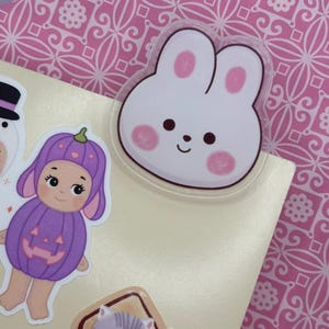 May include: A collection of colorful stickers featuring a white bunny with pink accents, a doll in a purple pumpkin costume, and a gray cat. The stickers are on a light-colored surface, with a pink patterned background.