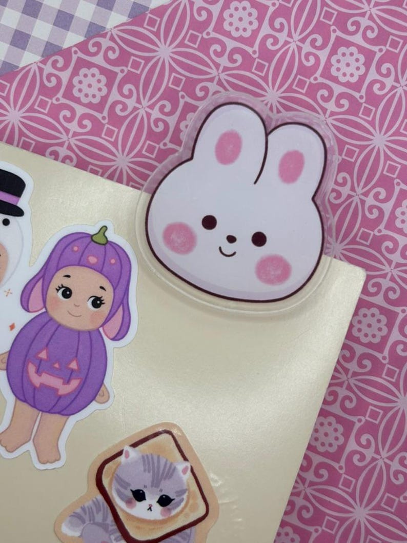 May include: A collection of colorful stickers featuring a white bunny with pink accents, a doll in a purple pumpkin costume, and a gray cat. The stickers are on a light-colored surface, with a pink patterned background.