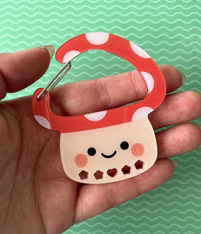 May include: A red and white polka-dotted mushroom-shaped carabiner clip. The clip has a smiling face with rosy cheeks and heart-shaped cutouts at the bottom. The clip is held in a hand.