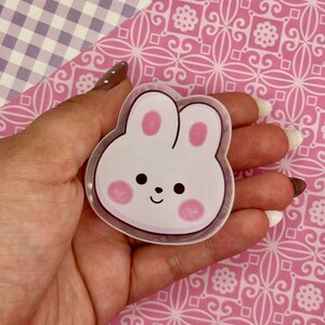 May include: A clear, bunny-shaped sticker with a white base, pink cheeks, and a small black smile. The sticker is held against a pink patterned background.