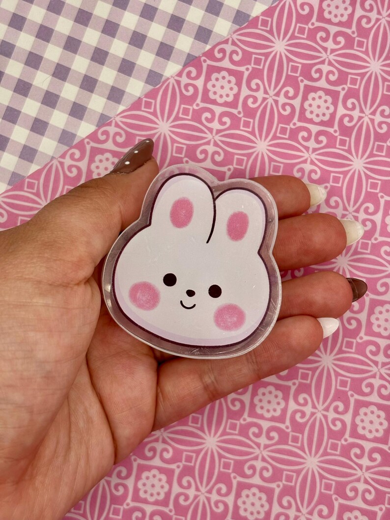 May include: A clear, bunny-shaped sticker with a white base, pink cheeks, and a small black smile. The sticker is held against a pink patterned background.