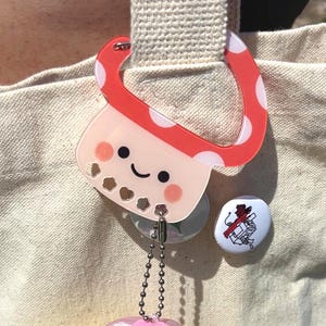 May include: A clear acrylic charm in the shape of a mushroom boba tea cup with a red and white polka dot top. The charm has a smiling face and is attached to a bag. Two small pins are also attached.