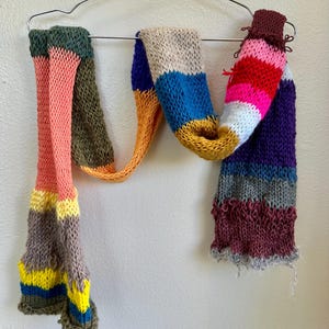 May include: A colorful, knitted scarf with a striped pattern. The scarf features a variety of colors, including pink, green, blue, yellow, and purple. It is displayed on a metal hanger against a white wall.