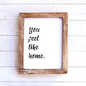 May include: A framed print with the words "You feel like home" in a black, handwritten-style font. The frame is made of light brown wood, and the background is white.