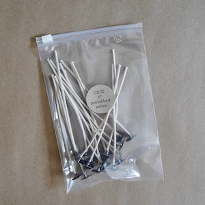 May include: A clear plastic bag filled with 22 white pre-tabbed candle wicks, each measuring 6 inches in length. The bag has a label that reads "CD 22 6" pretabbed wicks".