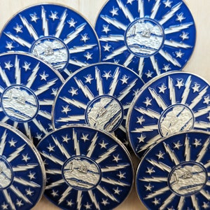 Può includere: Primo piano di otto spille in smalto blu e argento con un design a stella bianca e una nave bianca al centro. Le spille sono disposte in cerchio su una superficie di legno.