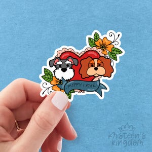 Puede incluir: Una pegatina con dos perros de dibujos animados dentro de un corazón rojo con una pancarta que dice "Puppy Love". La pegatina tiene flores naranjas y hojas verdes. El fondo es azul claro.