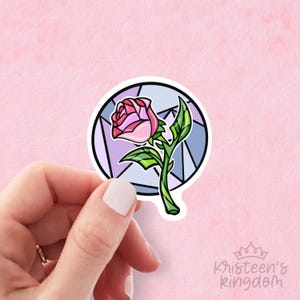 Puede incluir: Una pegatina con una rosa rosa con hojas y tallo verdes, enmarcada por un círculo con efecto vidriera. El diseño tiene un contorno negro y lo sostiene una mano sobre un fondo rosa. El texto "Kristeen's Kingdom" está en la esquina inferior derecha.