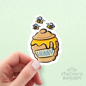 May include: A sticker featuring a cartoon honey pot with the word "HUNNY" in blue. Three yellow and black bees with blue wings are above the honey pot. The sticker is held by a hand against a light green background.
