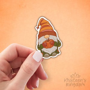 May include: A sticker of a cartoon gnome holding a pumpkin. The gnome is dressed in a striped orange and brown hat, a green sweater, and grey trousers. The sticker has a white border and is held by a hand against an orange background.