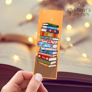 Puede incluir: Marcapáginas naranja con una pila de libros ilustrados con cubiertas coloridas. El marcapáginas tiene una base marrón y un fondo naranja claro con pequeñas estrellas blancas. El texto "Kristeen's Kingdom" está en la parte superior derecha.