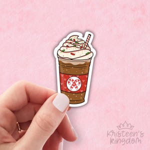 Puede incluir: Una pegatina de una bebida de café festiva con crema batida, espolvoreo de bastón de caramelo y un diseño de bastón de caramelo rojo y blanco en la taza.