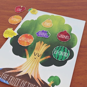 May include: A colorful illustration of a tree with a smiling face, featuring various fruits labeled with words like "Love," "Joy," and "Patience." The artwork is on a white background with a wooden table.