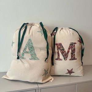 May include: Two off-white drawstring bags made of fabric, each featuring a large letter appliqué and scattered star motifs. The larger bag displays the letter A in a green floral pattern, while the smaller bag features the letter M in a red floral pattern. Both bags have dark green ribbon drawstrings.