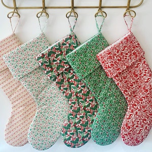 May include: Five Christmas stockings with different patterns. The stockings are made of fabric and have a white cuff. The patterns include red and green holly, red and white snowflakes, and a red and white floral design.