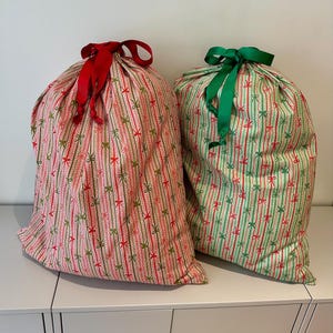 May include: Two fabric gift sacks featuring a vertical striped pattern with small bow motifs. The left sack has a light background with red and green bows and a red ribbon tie. The right sack has a light background with green and red bows and a green ribbon tie.