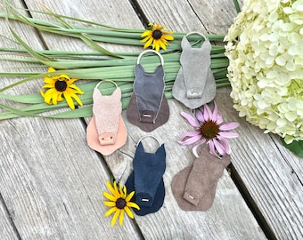 Peut inclure: Collection de six porte-clés en cuir en forme de tête de cheval, de différentes couleurs, dont rose, gris et bleu marine. Chaque porte-clés est orné d'un anneau métallique et est disposé sur une surface en bois avec des fleurs et de la verdure.