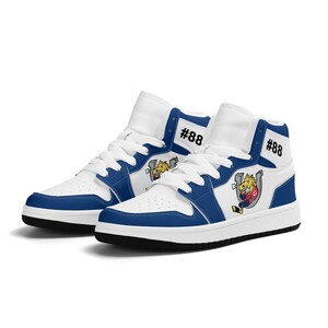 Puede incluir: Zapatillas blancas de caña alta con detalles azules y un logotipo de mascota de dibujos animados. Las zapatillas tienen una suela negra y el número #88 en el lateral.