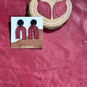 May include: A pair of burgundy arch earrings with black circular tops, displayed with two burgundy leaf-shaped earrings on a wooden slice. The earrings are on a white card with the text "111Craft". The background is a textured burgundy surface.