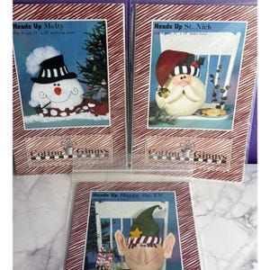 May include: Three craft pattern packets by Cotton Ginnys titled Heads Up Melty, Heads Up St. Nick, and Heads Up Happy the Elf. Each package features a photograph of a finished fabric character head, including a snowman, Santa, and an elf.