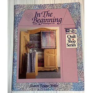 May include: A softcover book titled In The Beginning, Seattle, Washington USA, from the Quilt Shop Series by Sharon Evans Yenter. The cover features a light purple border with a blue and white speckled pattern and a wooden armoire filled with fabric.