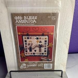 May include: A craft pattern packet titled God Bless America designed by Diane Arthurs from The Powder Mill. The cover features a red background with a quilt illustration showing two figures, stars, and hearts, enclosed in a clear plastic sleeve.